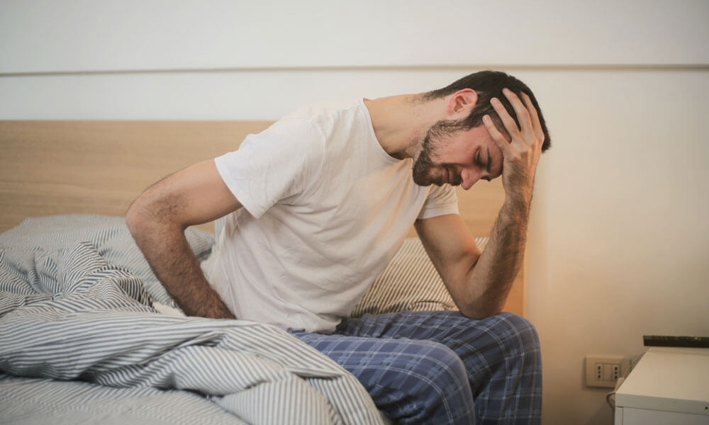 Slika prikazuje moškega, ki je vstal z postelje in se drži za glavo zaradi migrene.